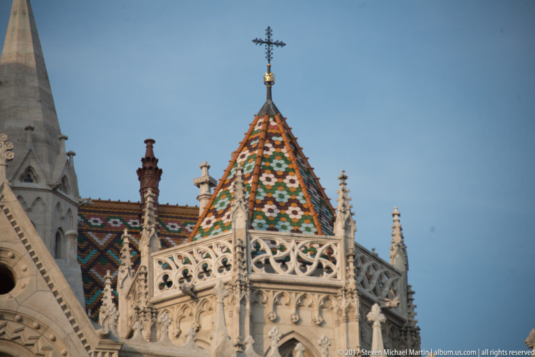 Decorative patterned roof tiles, carved stone balustrade, crocketed pinnacles, and a cross on Matthias Church.