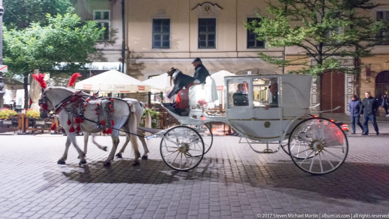 White horse-drawn carriage with two decorated gray horses on a cobbled Krakow square, with cafés and pedestrians behind.