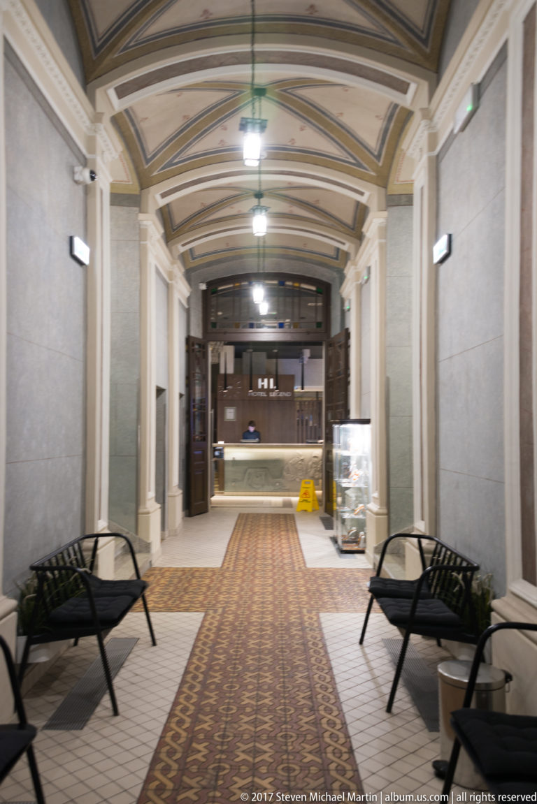 Vaulted corridor with patterned tile floor, black benches, wall lights, and a reception counter at the far end.