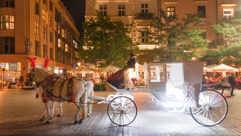 Brightly illuminated white horse-drawn carriage with two decorated horses and driver in Krakow’s Old Town at night.