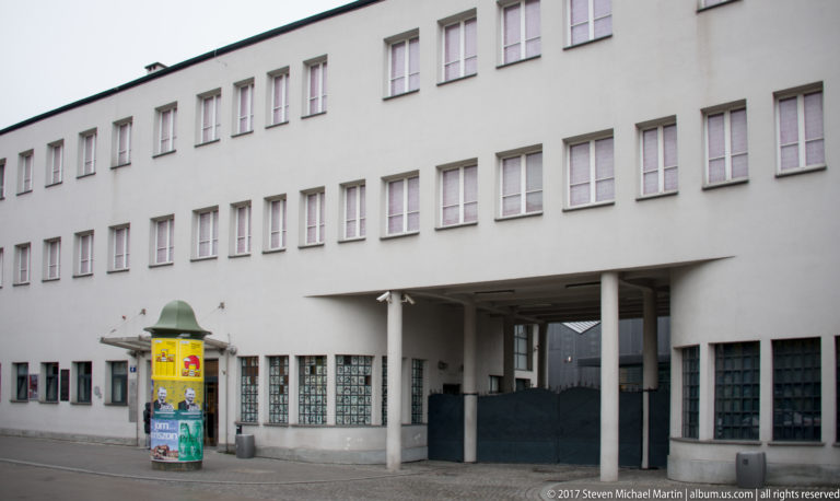 White exterior of Oskar Schindler’s former enamel factory in Kraków, with repetitive windows, recessed entrance, and advertising column.