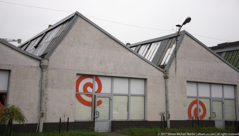 Low industrial building near Schindler’s Factory with sawtooth roofs, roof glazing, tall doors, and large red circular graphics.