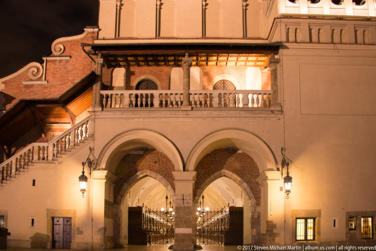 Twin stone arches with wrought gates beneath a balustraded staircase and brick vaulting in a historic Krakow building.