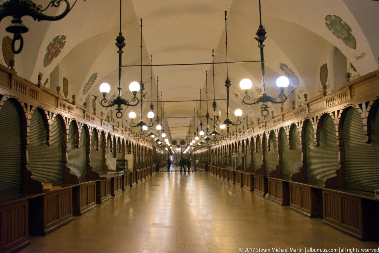 Symmetrical interior passage of the Krakow Cloth Hall with shuttered market stalls, hanging lamps, arches, and heraldic decoration.
