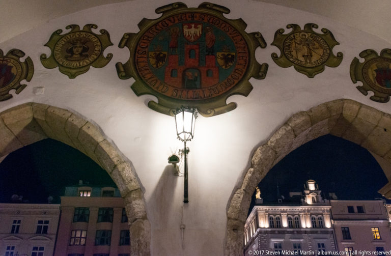 Painted heraldic shields above pointed arches and a lantern on the exterior arcade of Krakow’s Cloth Hall at night.