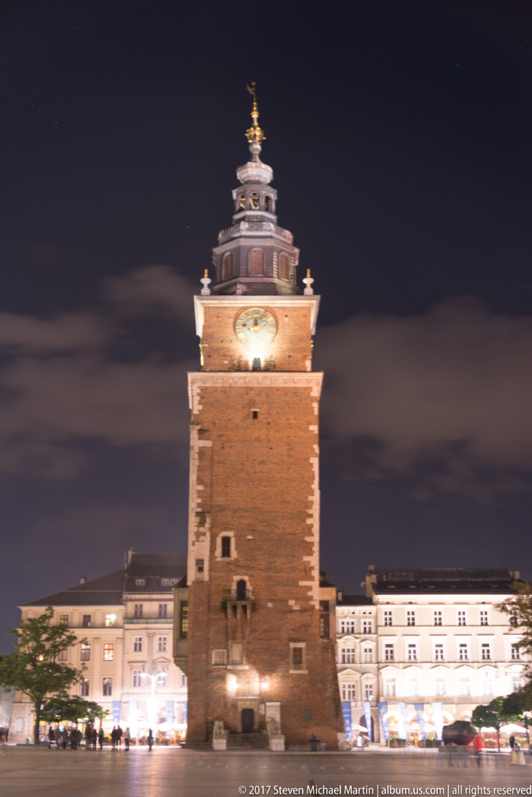 Krakow Town Hall Tower illuminated at night, with its brick shaft, clock, and baroque crown above the market square.