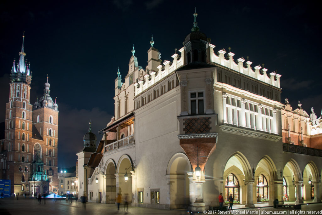Corner of Krakow’s illuminated Cloth Hall with pointed arcades, while St. Mary’s Basilica towers rise behind at night.