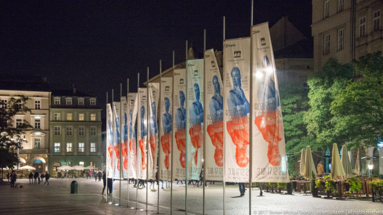 Row of tall portrait banners in red and white along a Krakow square at night, with cafés and historic buildings behind.