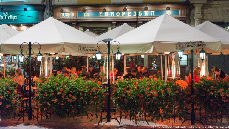 Evening terrace of Restaurant Europejska with white umbrellas, red flower boxes, diners, and outdoor heaters in Krakow.