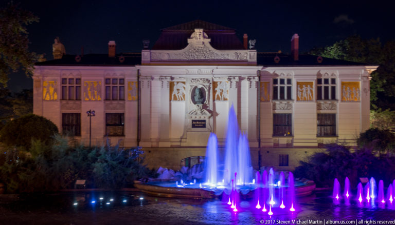 Color-lit fountain jets in Szczepański Square before the ornate illuminated façade of Krakow’s Palace of Art at night.