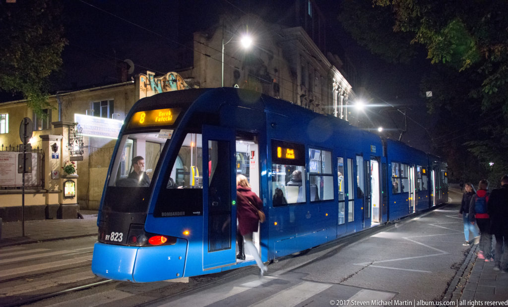 Blue Krakow tram stopped at night as passengers board beneath overhead electric wires in the historic city center.