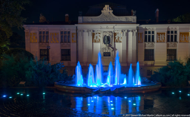 Blue illuminated fountain jets reflected on wet paving, with Krakow’s Palace of Art centered behind them at night.