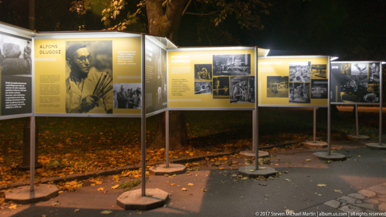 Outdoor exhibition panels at night, including a prominent display on Alfons Długosz with portrait and archival photographs.
