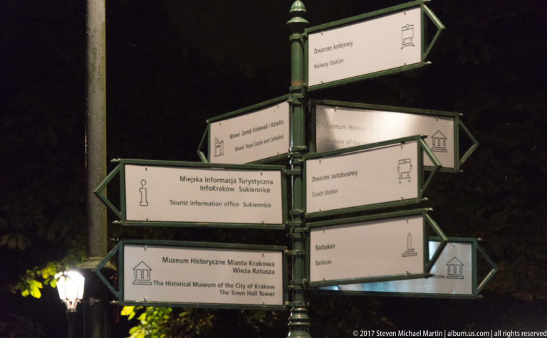 Green-and-white bilingual tourist signpost at night pointing to Krakow landmarks including the Barbican and Town Hall Tower.
