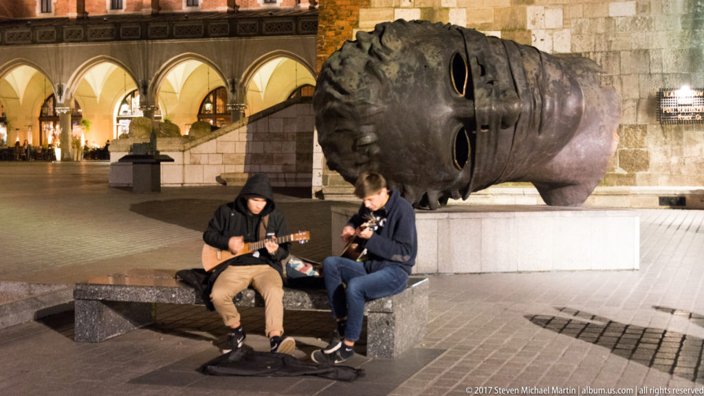 Two street musicians sit beside Igor Mitoraj’s giant bronze head sculpture Eros Bendato on Krakow’s Main Market Square at night.
