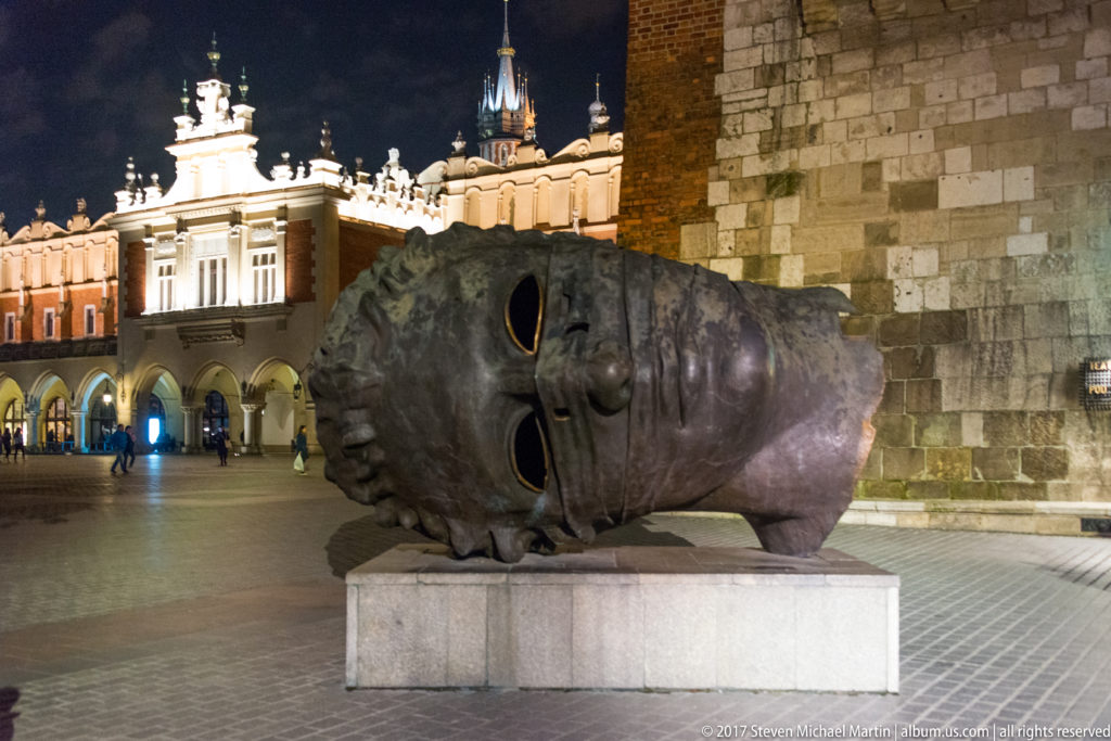 Close night view of Igor Mitoraj’s monumental bronze head Eros Bendato on a plinth beside Krakow’s Town Hall Tower.