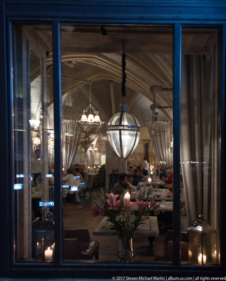 Restaurant dining room seen through a window at night, with spherical glass pendant, candles, curtains, and pink flowers.