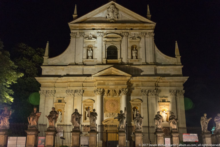 Floodlit Baroque façade of Krakow’s Church of Saints Peter and Paul behind a row of stone apostle statues at night.
