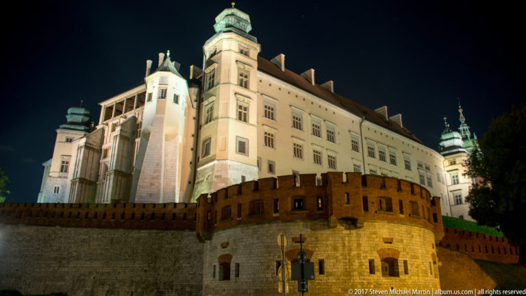 Illuminated Wawel Royal Castle above brick and stone defensive walls at night, viewed from below on Wawel Hill.