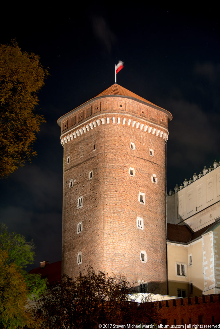Round brick defensive tower illuminated at night in Kraków, with a Polish flag above the roofline.