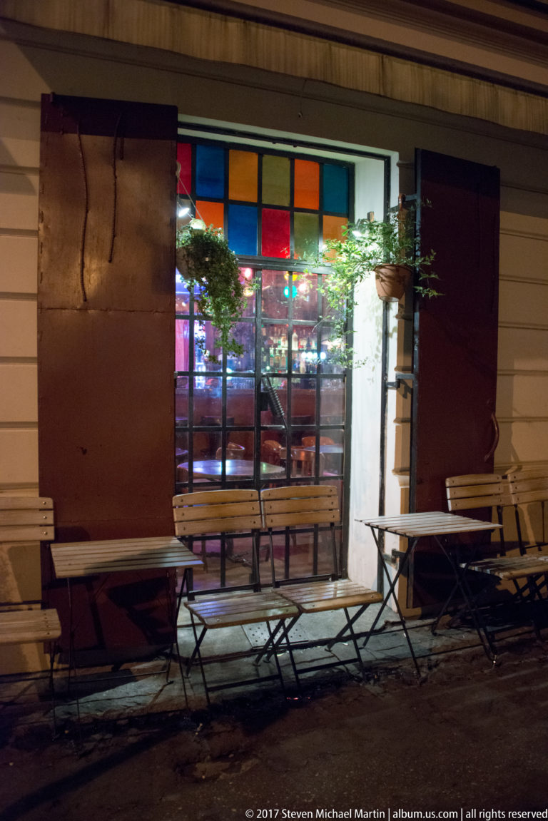 Nighttime café entrance with colorful transom panes, potted plants, and empty outdoor tables and chairs in Kraków.