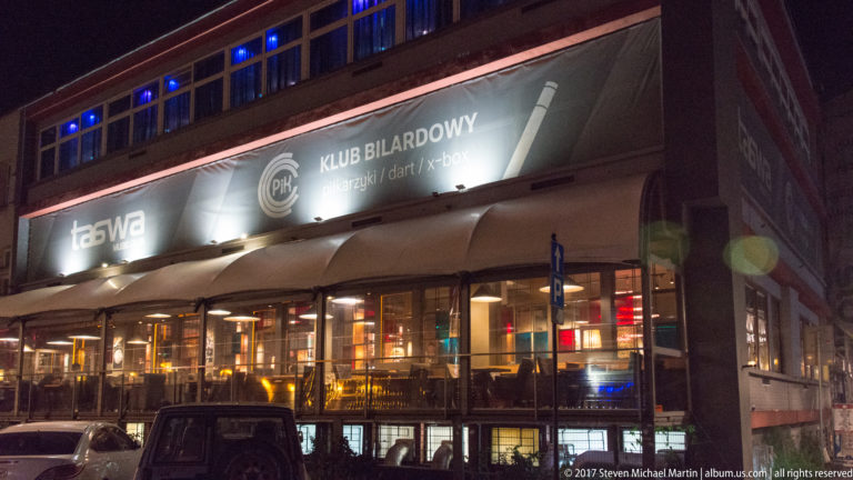 Night exterior of a Kraków entertainment venue with illuminated Taboo and billiards-club signage above awnings and large windows.