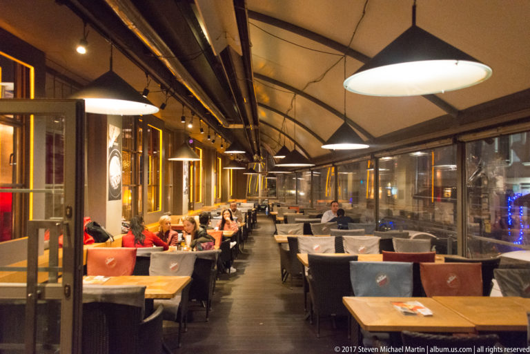 Covered restaurant terrace in Kraków with large hanging lamps, transparent side panels, diners, and rows of tables.