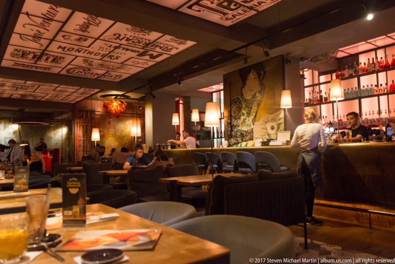 Warmly lit Kraków restaurant interior with bar shelves, diners, upholstered seating, decorative ceiling panels, and wall art.