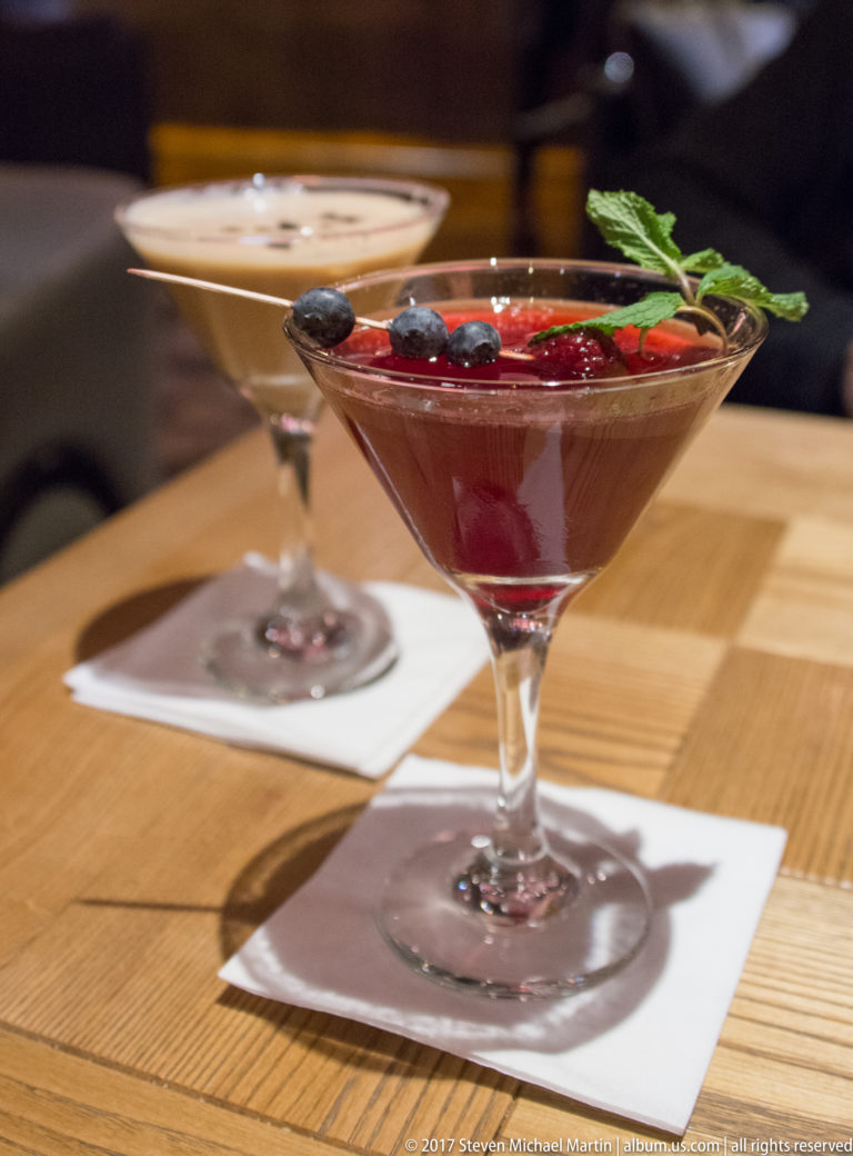 Deep red cocktail in a stemmed glass, garnished with berries and green herbs on a restaurant table.