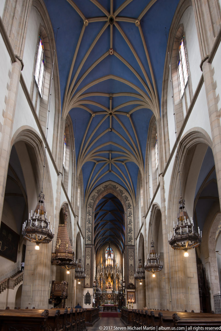 Wide Gothic church nave in Kraków with blue rib vaults, tall stone piers, chandeliers, and an illuminated high altar.