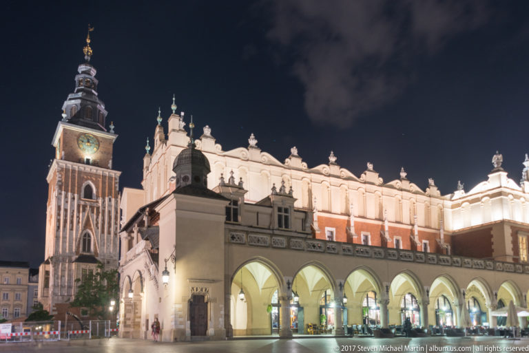 Illuminated Cloth Hall arcades and ornate roofline beside Krakow’s brick Town Hall Tower at night.