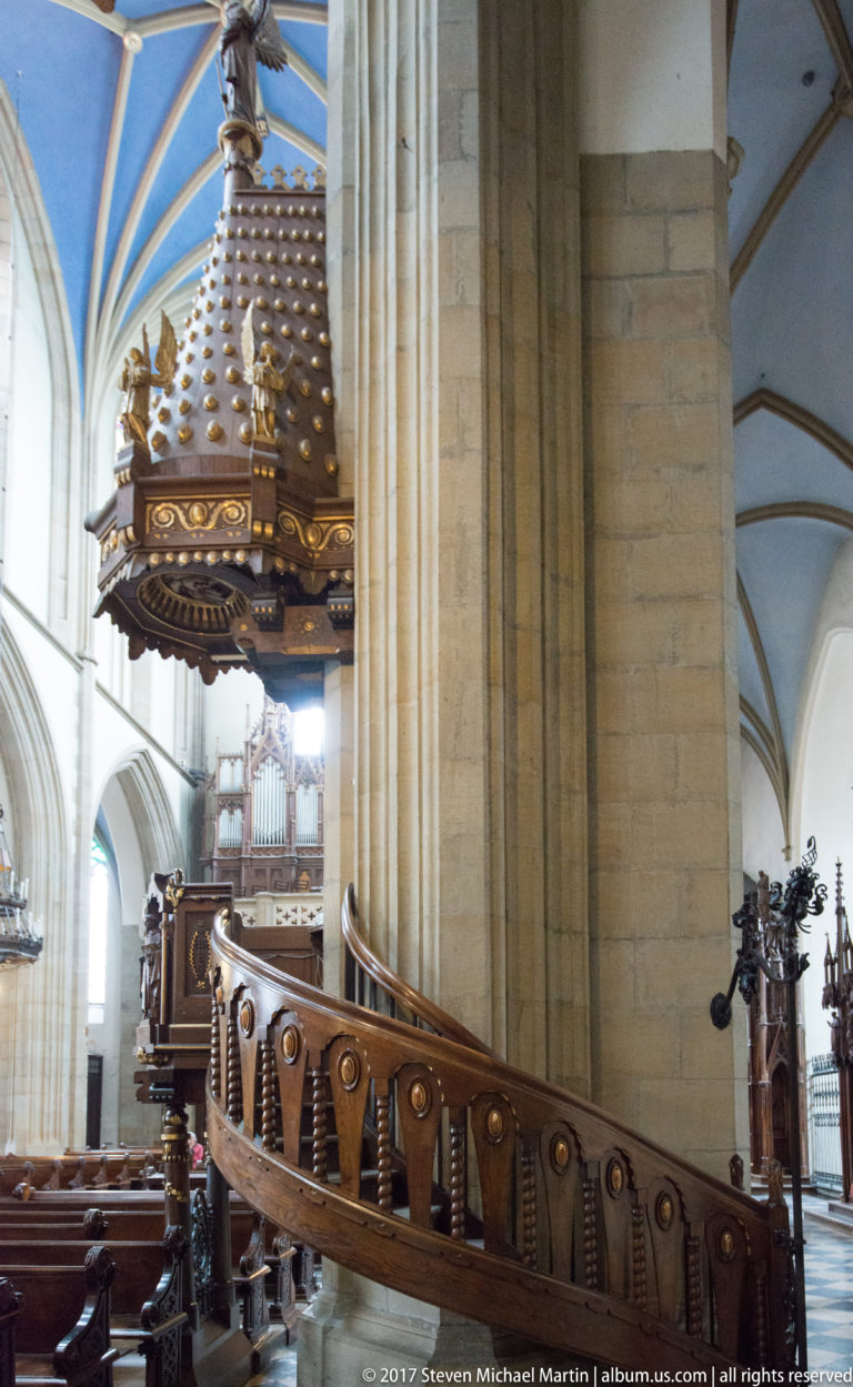 Carved wooden stair and choir furnishings wrapping around a massive stone pier inside a Gothic church in Kraków.