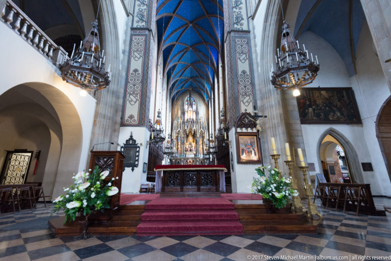 Frontal church sanctuary in Kraków with carved high altar, blue chancel vault, red steps, flowers, and patterned floor.
