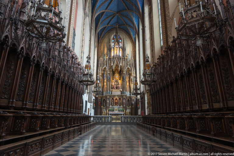 Long rows of carved wooden choir stalls leading toward a high altar beneath blue vaulting in a Kraków church.
