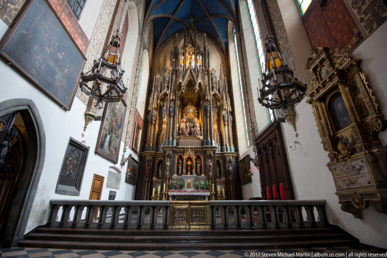 Ornate high altar beneath blue vaulting in a Kraków church, with side monuments, paintings, chandeliers, and stone balustrade.