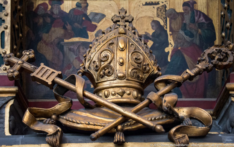 Ornate ecclesiastical emblem with bishop’s mitre, crossed ceremonial implements, drapery, and metalwork decoration in a Kraków church.