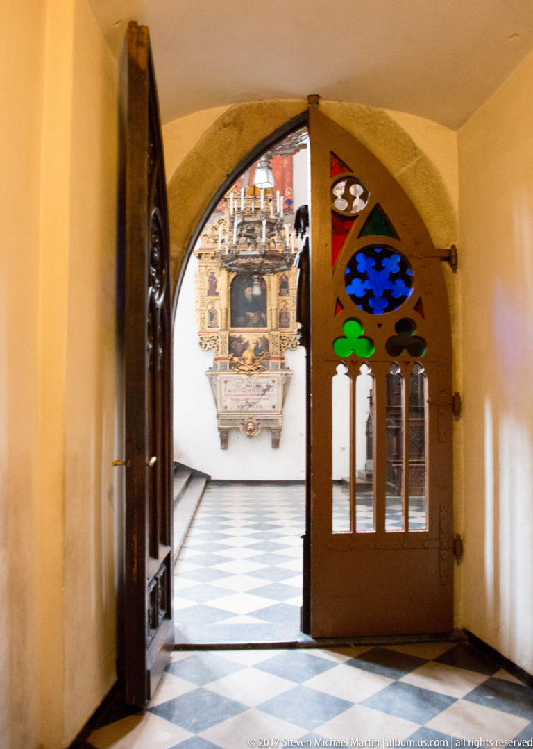 Open arched wooden doorway with colored-glass panels leading into a church interior with checker-pattern floor and ornate wall monument.