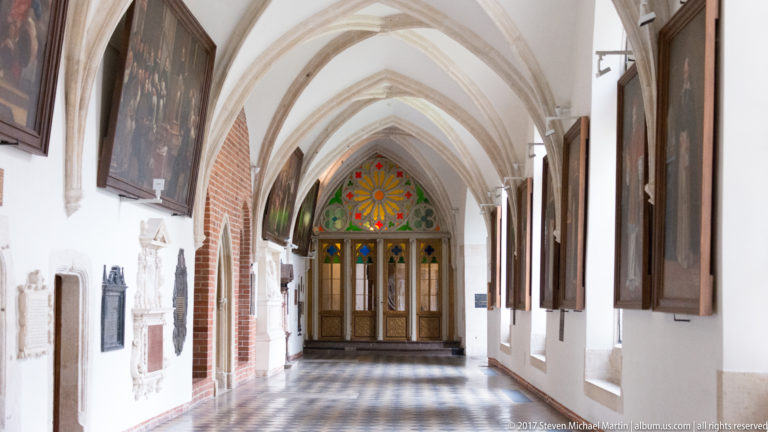 Vaulted church or monastic corridor in Kraków with brick arches, memorial plaques, paintings, patterned floor, and stained-glass doorway.