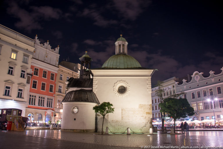 Small domed Church of St. Adalbert illuminated at night on Krakow’s Main Market Square, with historic façades behind.