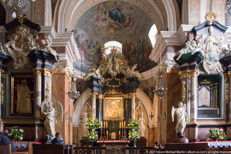 Ornate Baroque church sanctuary in Kraków with gilded altar, white sculptures, painted vault, side altars, and worshippers.