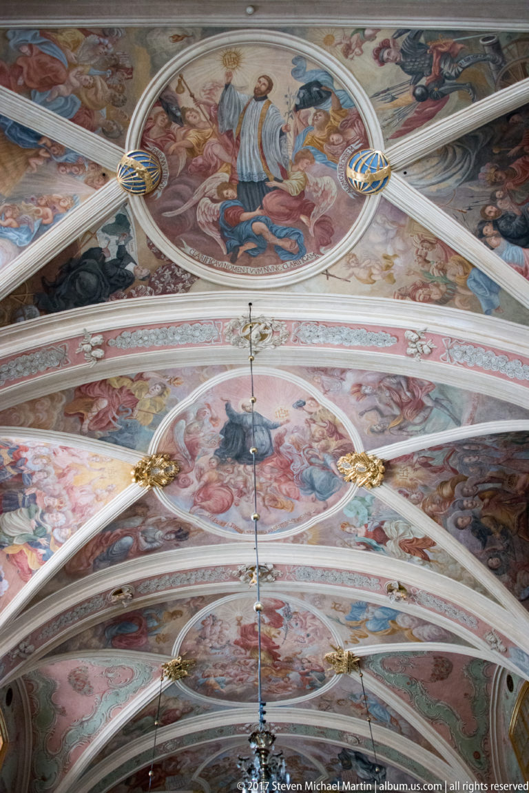 Painted vaulted church ceiling in Kraków with religious frescoes, pale ribs, gold rosettes, and a hanging central fixture.