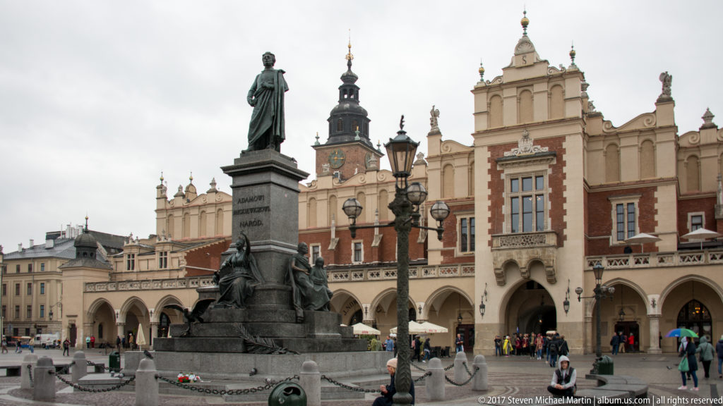 Adam Mickiewicz Monument in Kraków’s Main Market Square with the arcaded Cloth Hall behind it and pedestrians nearby.
