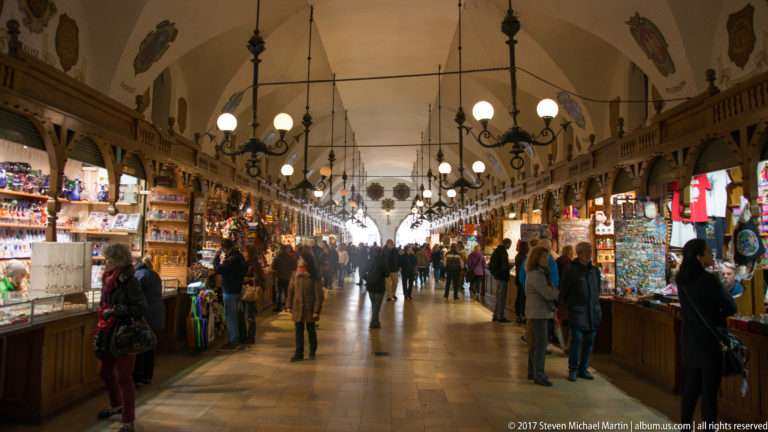 Interior of Kraków’s Cloth Hall with souvenir stalls, shoppers, repeating arches, chandeliers, and a long central aisle.