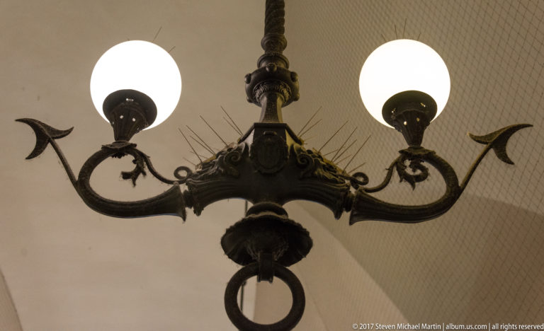 Decorative two-globe metal light fixture with curved pointed arms and central ring against a pale vaulted ceiling in Kraków’s Cloth Hall.
