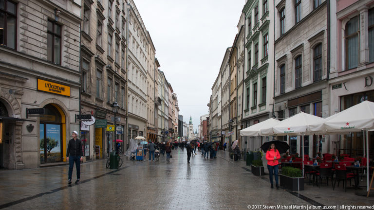 Wet pedestrian street in Kraków’s historic center with shops, café umbrellas, multi-story façades, and people walking.