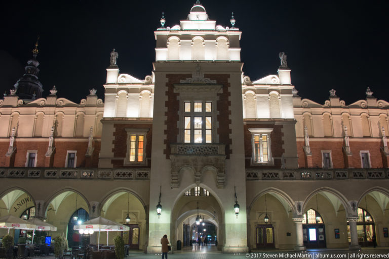 Floodlit central entrance of Krakow’s Cloth Hall with pointed arcades, red-brick panels, and ornate Renaissance parapet.