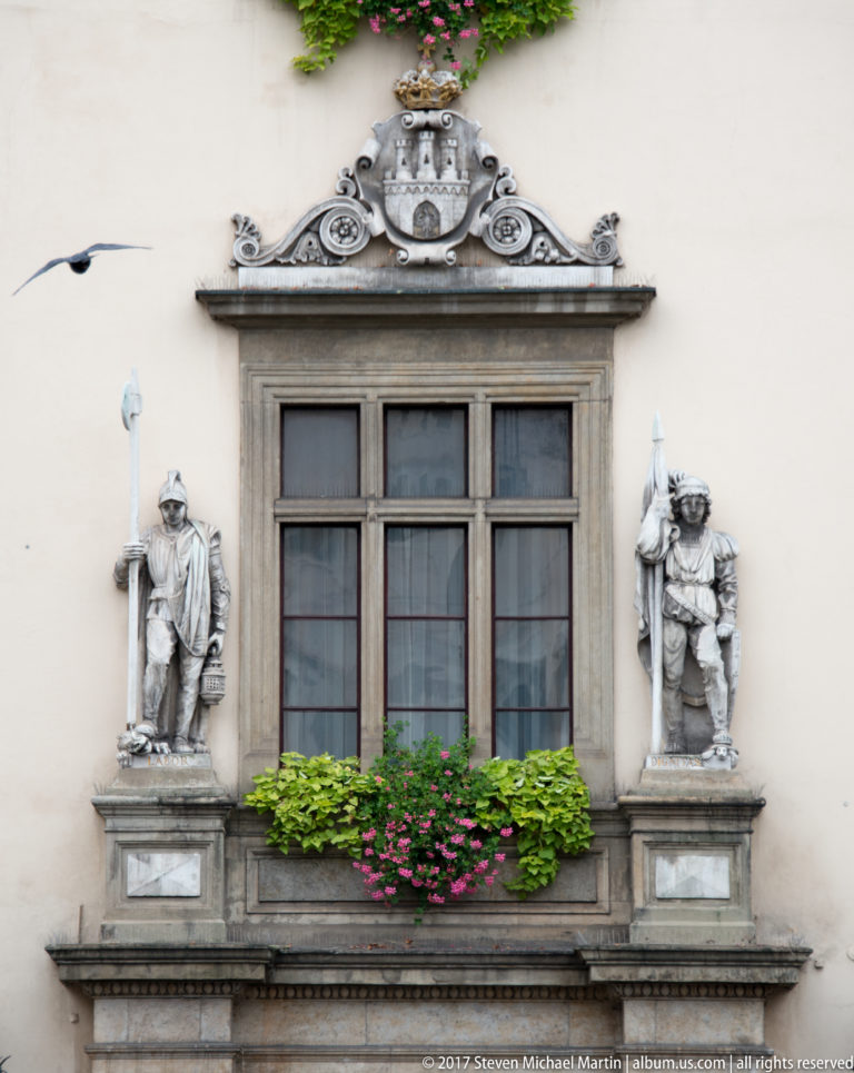 Historic Kraków window framed by two sculpted male figures, scrollwork, a small heraldic crest, and flower boxes.