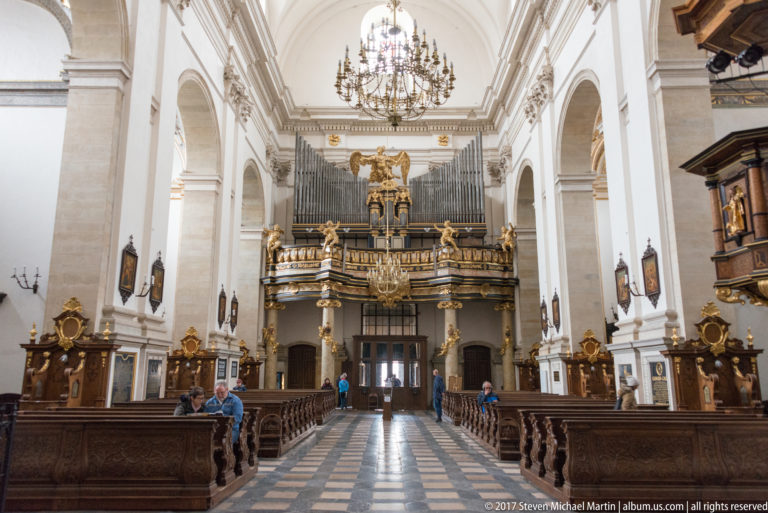 Bright Kraków church nave with dark carved organ loft, gilded ornament, chandeliers, white walls, and patterned central aisle.