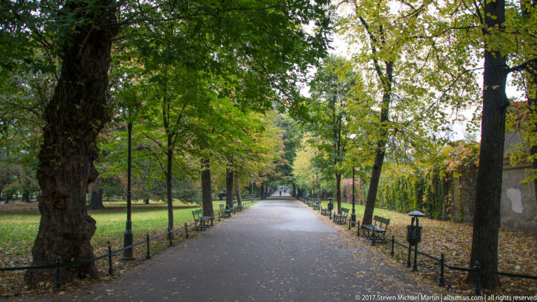 Tree-lined promenade in Kraków’s Planty Park with benches, lamps, railings, and fallen autumn leaves.