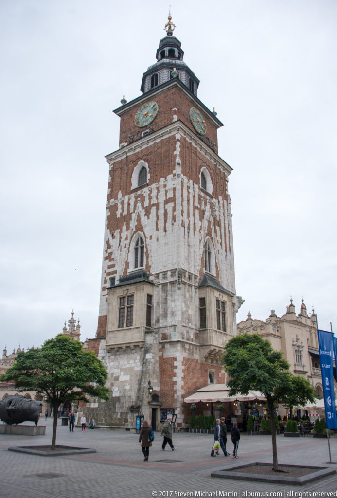 Town Hall Tower on Kraków’s Main Market Square, with brick walls, pale stone details, clocks, cupola, and pedestrians below.